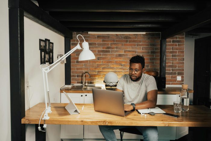 A man working from home at a desk with laptop, lamp, and workspace essentials.