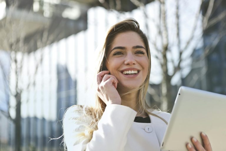 Confident businesswoman using her tablet and phone, smiling outdoors in sunlight.