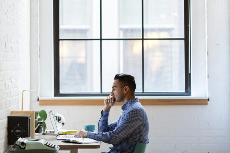 Focused male professional working on a laptop at an office desk by a large window.